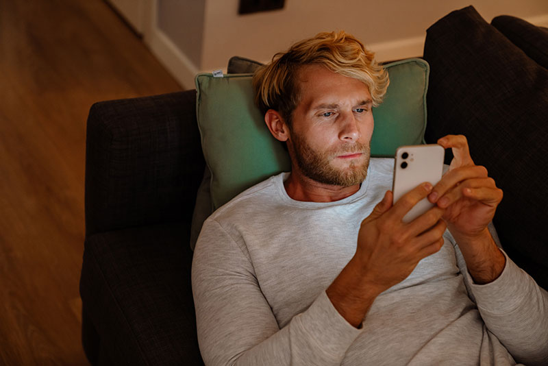 Blonder Mann liegt auf Couch und schaut auf sein Smartphone