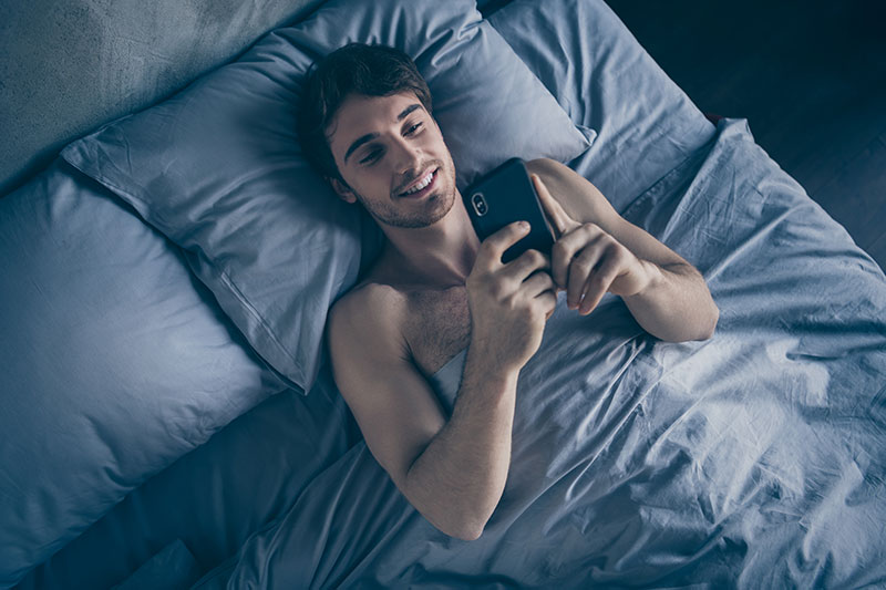 Junger Mann liegt im Bett und tippt am Handy